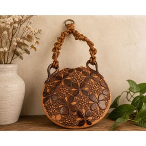 May include: A round, tan ceramic wall hanging featuring a dark brown, intricate lace-like floral pattern embossed on its surface. The piece includes a thick, braided brown rope handle attached to two curved ceramic loops at the top for hanging.
