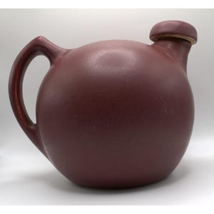 May include: A deep red ceramic pitcher with a round body and a single handle. The pitcher has a lid with a small knob on top.