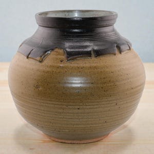 May include: A handcrafted ceramic vase with a rounded body and a dark brown rim. The vase has a textured, earthy brown finish with a decorative notched edge. The vase is a decorative item.