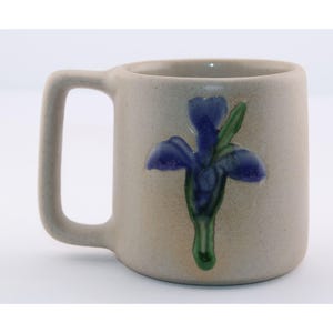 May include: A beige ceramic mug with a large handle. The mug features a blue iris flower design with green leaves. The mug is a neutral color and has a simple, classic design.