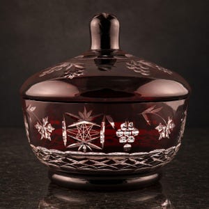 May include: A deep red glass bowl with a matching lid, featuring intricate cut-glass patterns including starbursts, grape clusters, and leaf motifs. The base and lid rim display a diamond-cut crosshatch design, showcasing clear glass accents.