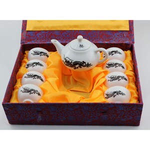 May include: A white ceramic tea set featuring a teapot and eight small cups, each decorated with a black dragon motif. The teapot lid displays a small black character. The set is presented in a rectangular box lined with bright yellow fabric.