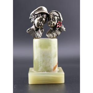 May include: A sculpture featuring two silver busts of children atop a green and beige onyx base. One child wears a cap, the other has a red flower in their hair. The base is rectangular and cylindrical.