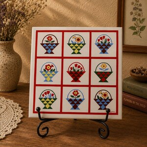 May include: A decorative square tile featuring a grid of nine squares, each with a basket design. The baskets are filled with colorful flowers and set against a white background with a red border. The tile is displayed on a black metal stand.