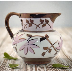 May include: A decorative ceramic pitcher with a brown handle and rim, featuring a white body adorned with pink floral and brown leaf designs. The pitcher has a spout for pouring and sits on a wooden surface.