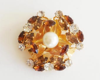 May include: A gold-toned brooch with a central pearl surrounded by brown and clear crystals. The brooch has a floral design with a layered appearance, creating a sparkling, elegant accessory.