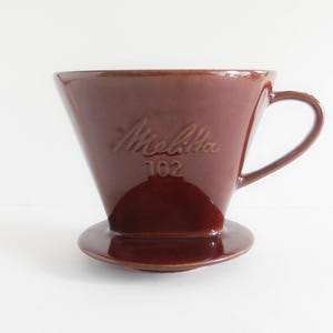 Pode incluir: Cafeteira Melitta 102 de cerâmica marrom, com alça e base pequena. A cafeteira em forma de cone tem o nome da marca e o número do modelo em relevo. Projetada para preparar café.