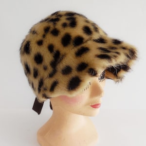 Peut inclure: Chapeau en fausse fourrure à imprimé léopard avec un bord incurvé et un ruban marron. Le chapeau est présenté sur une tête de mannequin. Les couleurs du chapeau sont le beige, le marron et le noir.