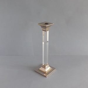 Può includere: Un singolo portacandele con base e parte superiore quadrate. La base e la parte superiore sono di colore oro metallizzato e lo stelo è in acrilico trasparente e rettangolare. Il candeliere è centrato su uno sfondo grigio.