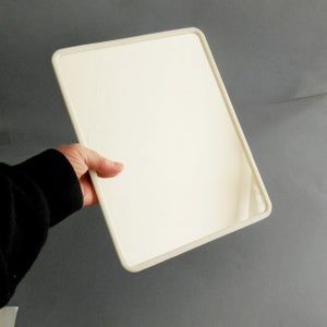 May include: A rectangular, off-white mirror with a rounded frame is held by a person wearing a black sweater. The mirror's surface is smooth and reflective, with a slight crack visible. The frame is a lighter shade of off-white.