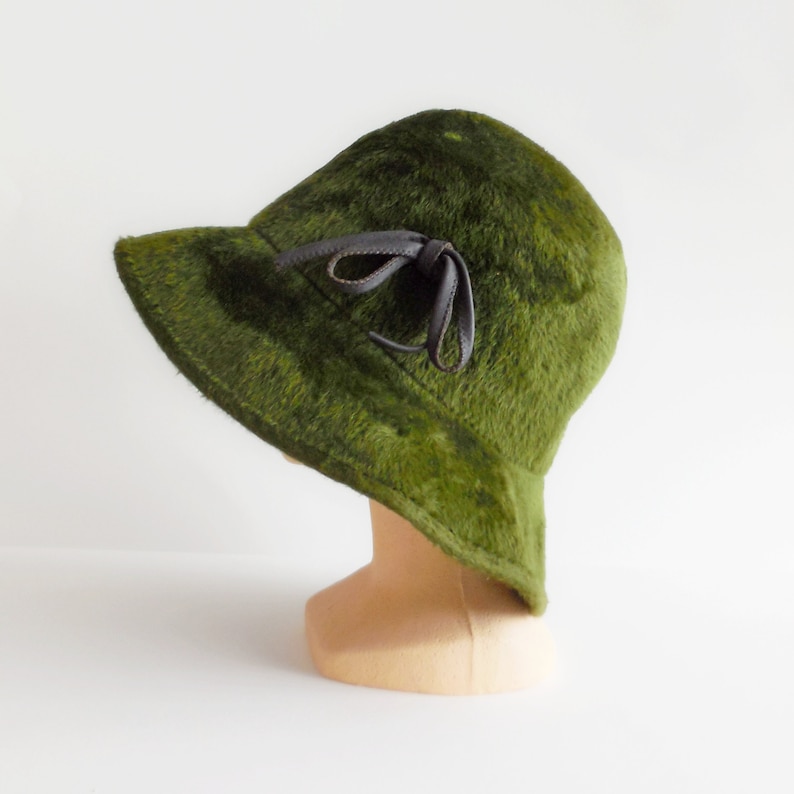 Peut inclure: Un chapeau vert vintage avec un nœud en cuir marron foncé. Le chapeau a un aspect doux et texturé et un bord incliné vers le bas. Le chapeau est présenté sur une tête de mannequin sur fond blanc.