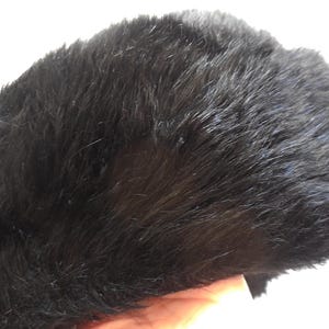 May include: A black faux fur hat. The hat has a dark, solid colour with a soft, textured appearance. The hat is shown against a white background.