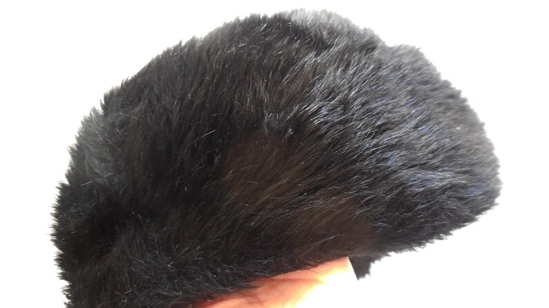 May include: A black faux fur hat. The hat has a dark, solid colour with a soft, textured appearance. The hat is shown against a white background.