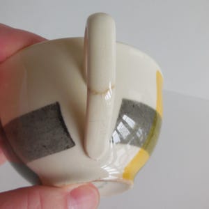 May include: Cream-colored ceramic teacup with a curved handle. The cup features a geometric design of black, gray, and yellow squares. The cup is held by a hand.