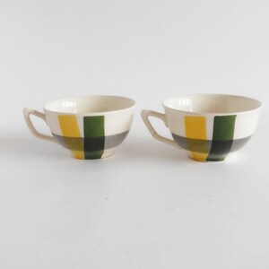 May include: Two white ceramic teacups with handles. Each cup features a geometric design of yellow, green, and gray squares. The cups are set against a white background.