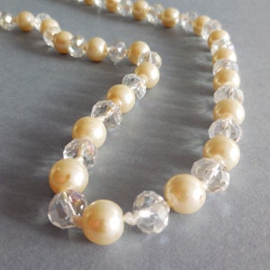May include: A cream-coloured necklace featuring alternating large, round faux pearls and clear, faceted crystal beads. The necklace is strung on a white cord against a light grey background.