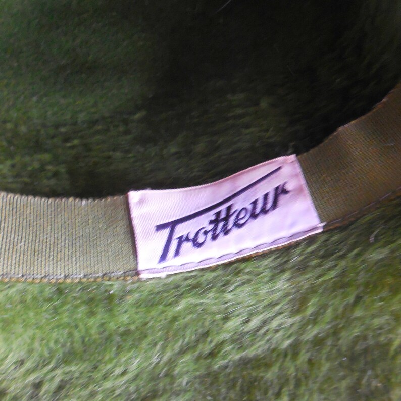 May include: Close-up of a green hat with a soft, textured surface. The interior features a light pink label with the word "Trotteur" in a dark blue script. The hat's brim is a darker green.
