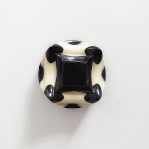 Puede incluir: Botón cuadrado negro con base crema y lunares negros. El botón tiene un centro cuadrado en relieve y probablemente se utiliza para ropa o manualidades.