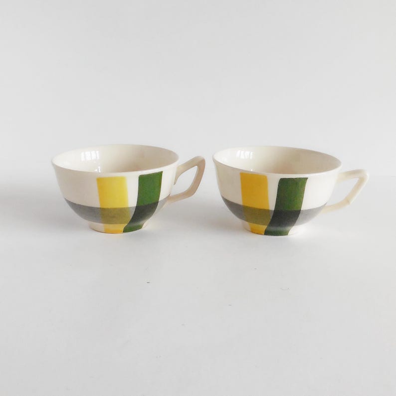 May include: Two white ceramic teacups with handles. Each cup features a design of yellow, green, and gray stripes. The cups are set against a white background.
