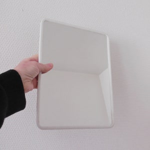 May include: A white, rectangular mirror with rounded corners is held by a person. The mirror has a clean, minimalist design and is set against a textured white wall. The person's arm and hand are visible, wearing a black sleeve.