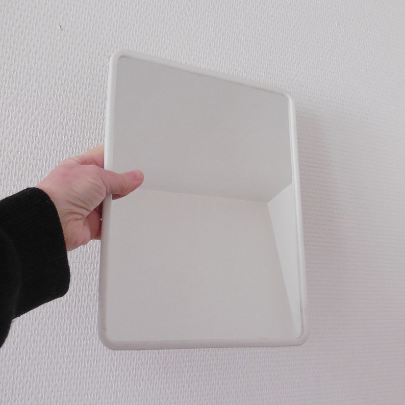 May include: A white, rectangular mirror with rounded corners is held by a person. The mirror has a clean, minimalist design and is set against a textured white wall. The person's arm and hand are visible, wearing a black sleeve.
