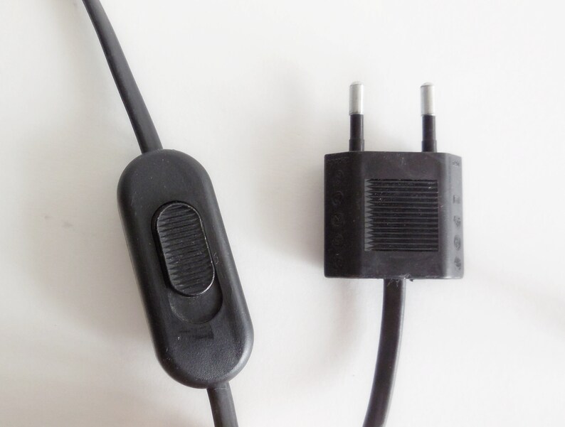 以下が含まれることがあります： 黒い電気コードのクローズアップ。オン/オフスイッチと2ピンのプラグが内蔵されています。スイッチは楕円形で、ボタンがくぼんでいます。プラグは長方形で、2つの銀色のピンが付いています。