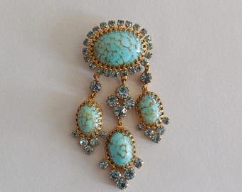May include: A gold-tone brooch featuring a large oval turquoise-colored cabochon with brown veining, surrounded by small light blue rhinestones. Three smaller oval turquoise-colored cabochons dangle from the base, each accented with light blue rhinestones.