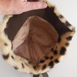 May include: A leopard print faux fur hat. The hat has a brown interior lining and a dark brown band around the brim. The fur is a mix of tan and cream colours with black spots. A brown ribbon is visible.