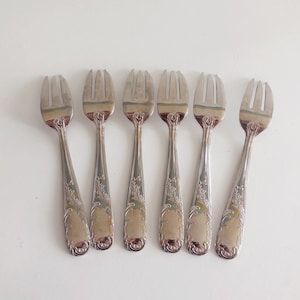 Peut inclure: Six fourchettes argentées avec des motifs floraux ornent les manches, disposées sur fond blanc. Chaque fourchette a trois dents et une finition brillante. Elles semblent être des couverts anciens ou vintage.