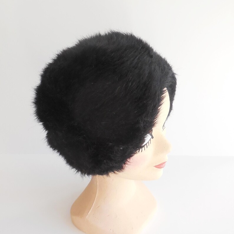 May include: A black, fluffy hat, possibly faux fur, is displayed on a mannequin head. The hat has a rounded shape and a dense, textured appearance. The mannequin head is a light beige colour, and the background is plain white.