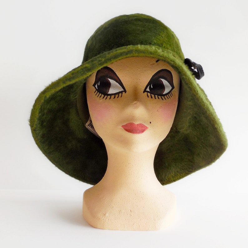 May include: A vintage green fur hat with a wide brim and a small black bow. The hat is displayed on a mannequin head with painted features, including large eyes and red lips. The hat's texture appears soft and plush.