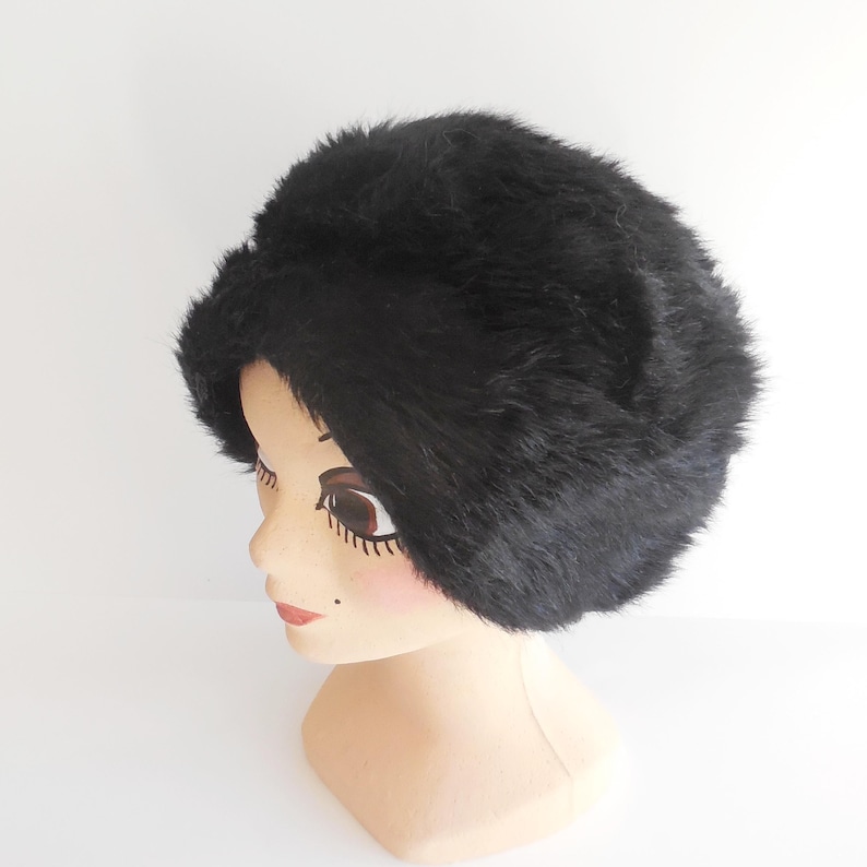 May include: A black faux fur hat with a rounded crown. The hat is displayed on a mannequin head with painted features, including brown eyes and red lips. The hat has a soft, textured appearance.