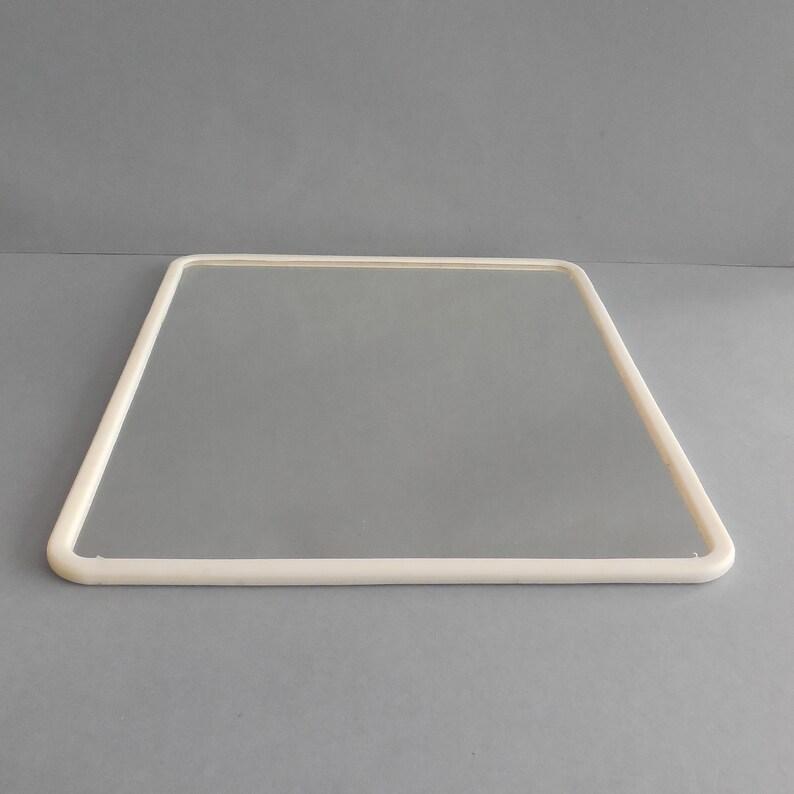 May include: A clear, rectangular glass pane with a rounded, off-white frame. The frame is a continuous, smooth line that outlines the glass. The image is set against a solid gray background, highlighting the simplicity of the design.