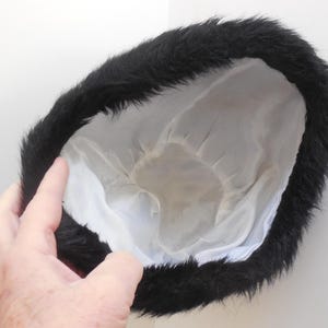 May include: A black fur hat with a white satin lining. The hat is round and has a fluffy, textured appearance. The hat is held by a hand against a white background.