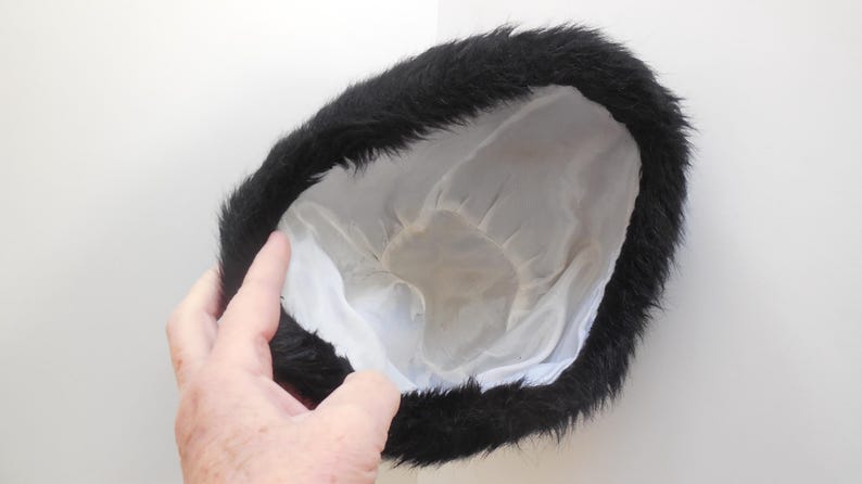 May include: A black fur hat with a white satin lining. The hat is round and has a fluffy, textured appearance. The hat is held by a hand against a white background.