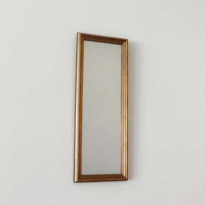 Op de afbeelding: Een rechthoekige spiegel met een goudkleurige lijst. De spiegel is lang en smal, met een eenvoudig, elegant ontwerp. De lijst heeft een licht getextureerde uitstraling. De spiegel is aan een witte muur bevestigd. Afmetingen: ongeveer 20 cm x 60 cm.