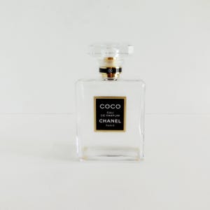 May include: A clear, rectangular perfume bottle with a clear, faceted cap. The bottle features a black label with gold lettering that reads "COCO Eau de Parfum CHANEL PARIS". A gold-coloured band encircles the neck.