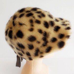 May include: A leopard print hat. The hat has a tan base colour with black spots, and a dark brown ribbon. The hat is made of a soft, faux fur material. The hat is a vintage style.
