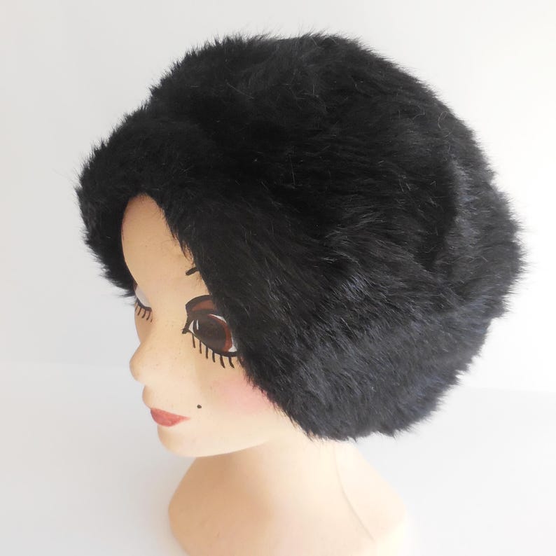 May include: A black faux fur hat. The hat is round and fluffy, with a soft texture. The hat is displayed on a mannequin head with painted features and a pale complexion. The background is plain white.