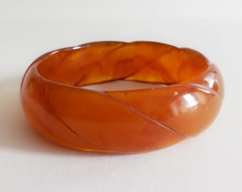 May include: A translucent amber-colored bangle bracelet featuring a smooth, polished surface with a repeating diagonal carved twist pattern that wraps around the entire circumference of the circular band, creating a continuous, flowing design.