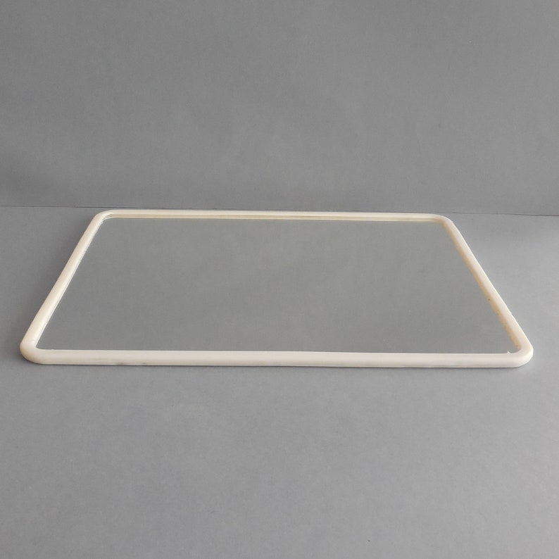 May include: A rectangular mirror with a rounded, off-white frame. The mirror is set against a gray backdrop, creating a clean and minimalist aesthetic. The frame's smooth lines and neutral color suggest a modern design.