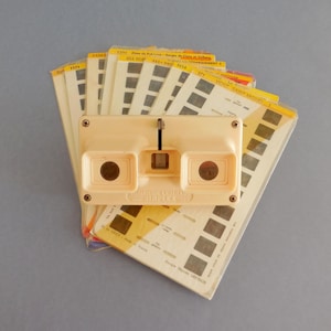 Peut inclure: Un ancien stéréoscope Simplex beige avec une pile de diapositives. Le viseur a deux trous pour les yeux et un réglage de la mise au point. Les diapositives sont rectangulaires avec des images et du texte en noir et blanc. Le fond est gris clair.