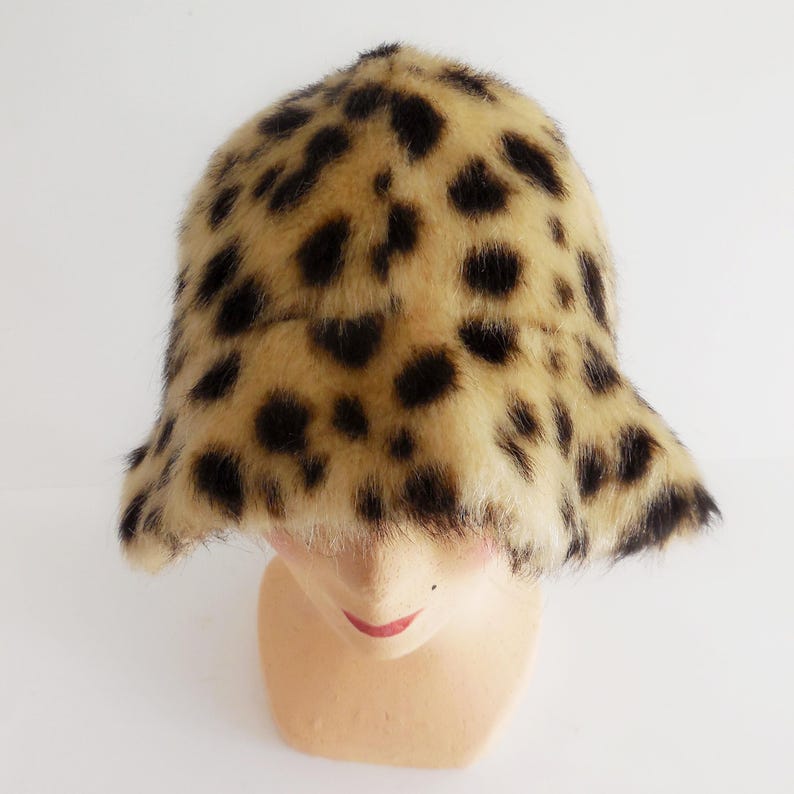 May include: A leopard print bucket hat. The hat has a tan base colour with dark brown spots. The brim is slightly flared. The hat is made of faux fur and is displayed on a mannequin head.