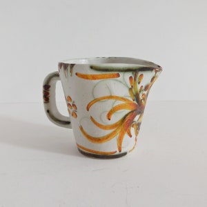 Può includere: Brocca in ceramica color crema con un motivo floreale decorativo nei toni dell'arancione, giallo e marrone. La brocca ha un manico e un beccuccio. Il motivo floreale è sul davanti e sul lato.