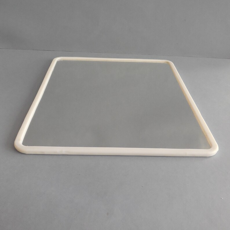 May include: A square, frameless mirror with a thick, off-white border. The mirror is rectangular with rounded corners and is set against a gray background. The border is smooth and uniform in width.