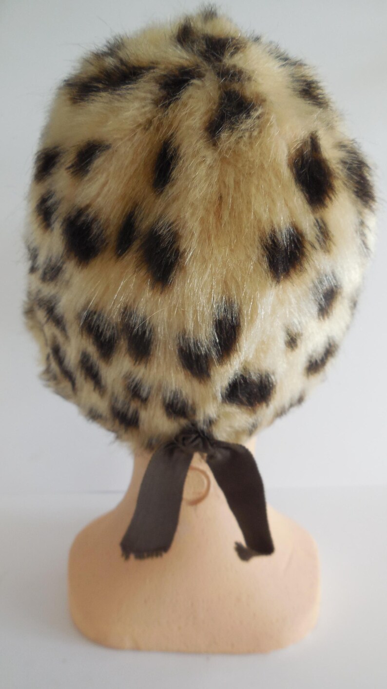 May include: A vintage-style hat with a faux fur leopard print design. The hat features a beige base with dark brown spots and a brown ribbon tie at the back. This accessory is a stylish addition to any wardrobe.