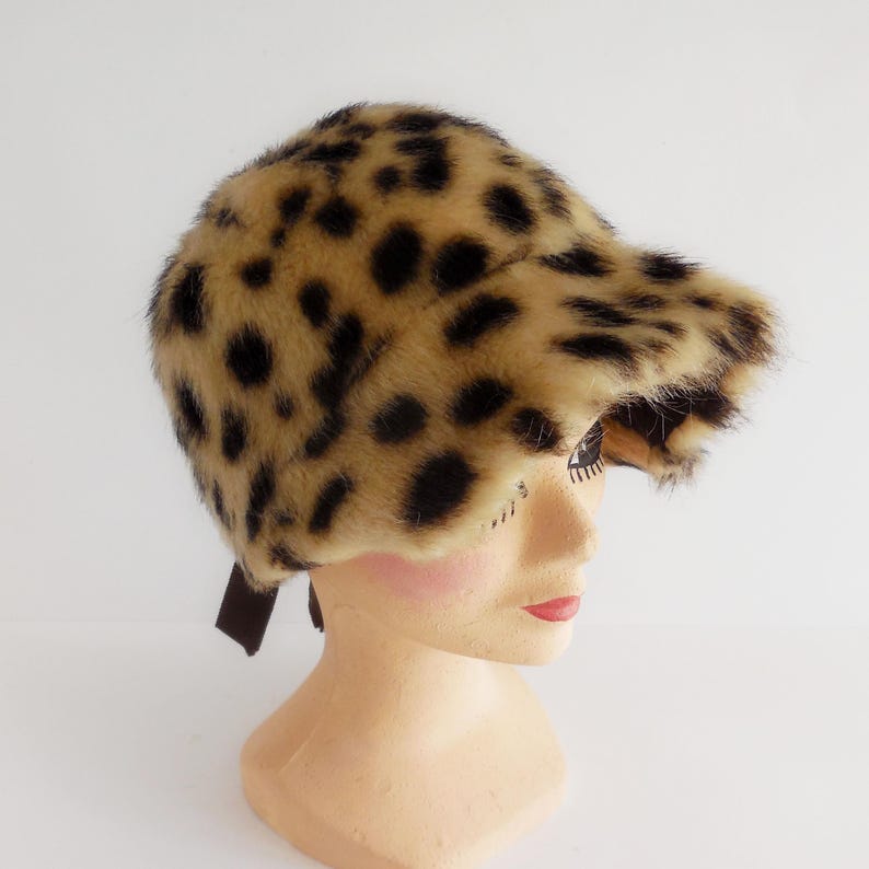 Peut inclure: Chapeau en fausse fourrure à imprimé léopard avec un bord incurvé et un ruban marron. Le chapeau est présenté sur une tête de mannequin. Les couleurs du chapeau sont le beige, le marron et le noir.