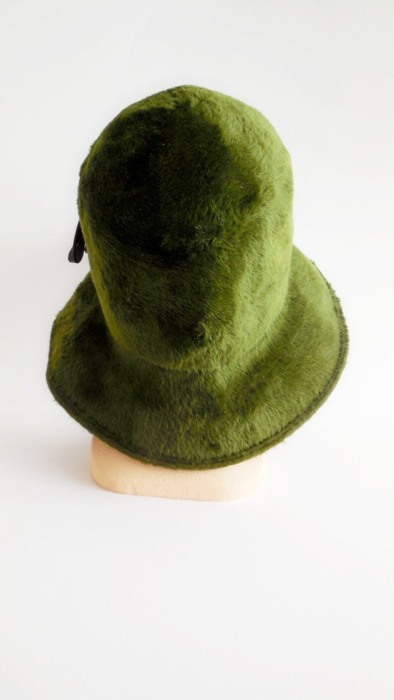 May include: A green fuzzy hat with a rounded crown and a wide brim. The hat is a solid, deep green color and has a soft, textured appearance. A black ribbon is attached to the back of the hat. The hat is displayed on a beige mannequin head.