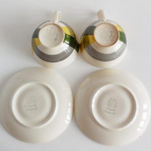 May include: Two teacups and saucers. The teacups have a striped pattern in yellow, green, and gray. The saucers are plain white. The teacups have a small handle. The saucers have a small, circular indentation in the center.
