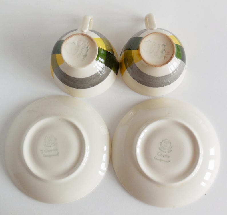 May include: Two teacups and saucers. The teacups have a striped pattern in yellow, green, and gray. The saucers are plain white. The teacups have a small handle. The saucers have a small, circular indentation in the center.